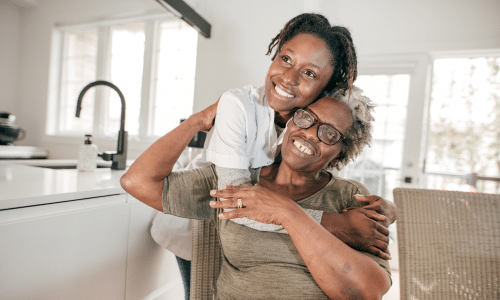 A young Black woman wearing a white top affectionately hugging an older Black woman wearing glasses and an olive shirt from behind in a bright, modern kitchen.