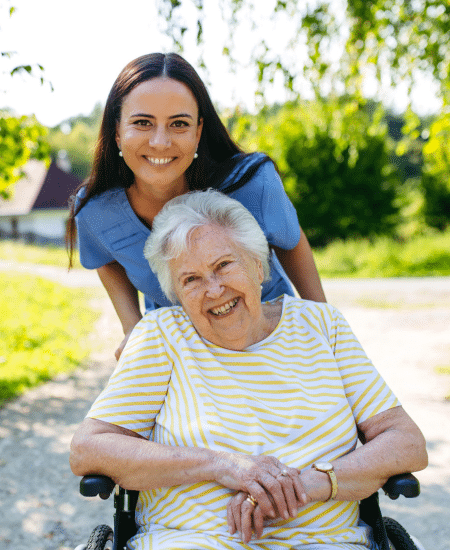 A young female caregiver smiling and pushing an elderly happy woman in a wheelchair outdoors on a sunny day.